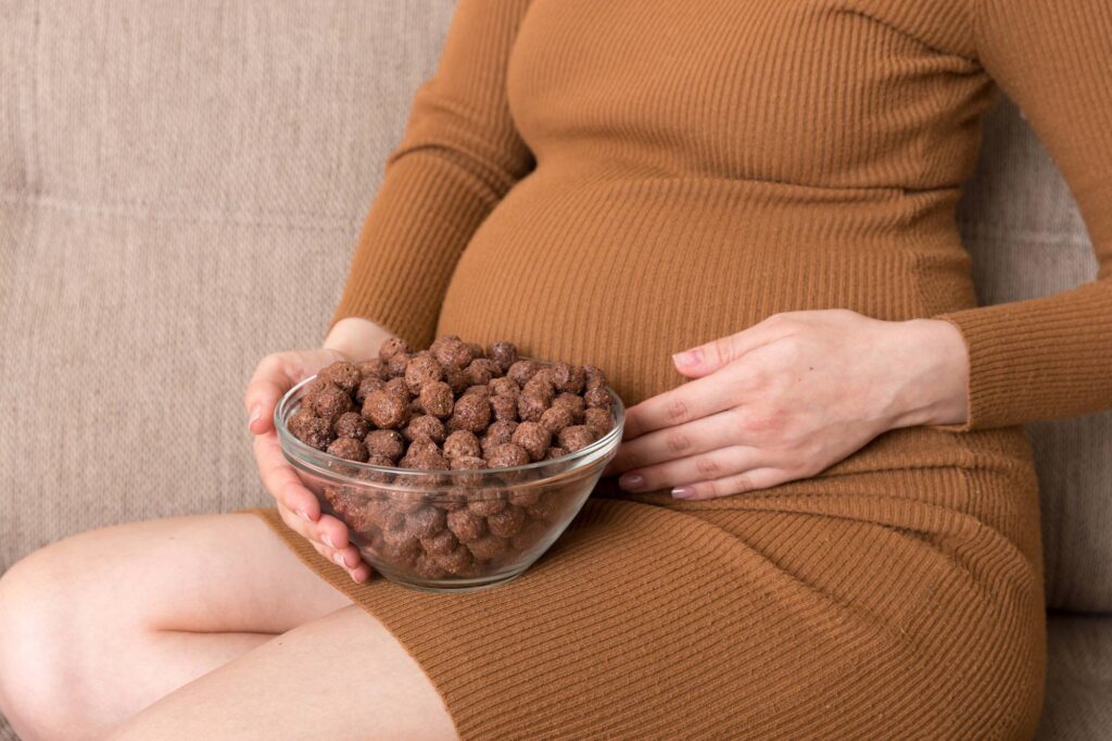 Close up of pregnant woman enjoys eating crunchy chocolate cereal balls from a bowl relaxing on the sofa. Dieting during pregnancy concept.