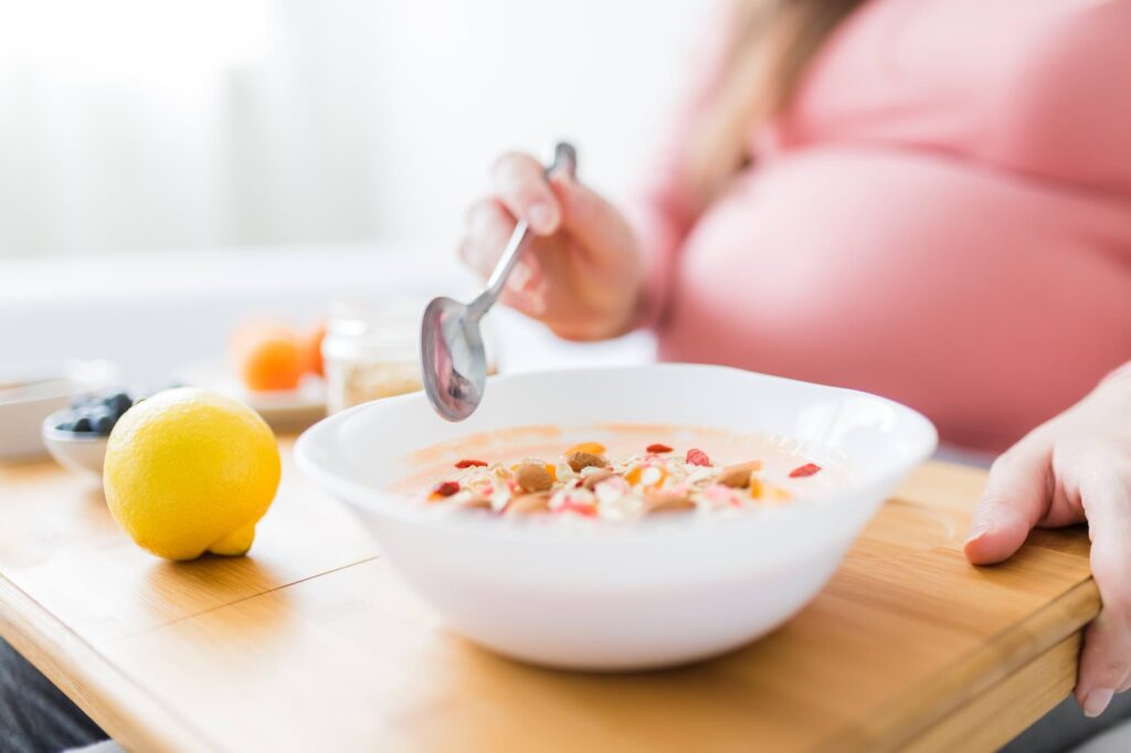 Close up of young pregnant woman sitting on bed and eating granola with cereals and yogurt.
